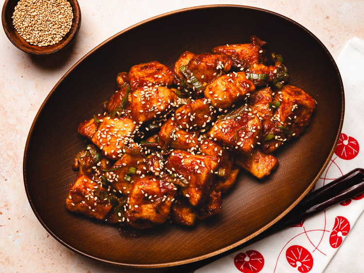 sweet and sour tofu in a bowl with a pinch bowl of sesame seeds in the corner